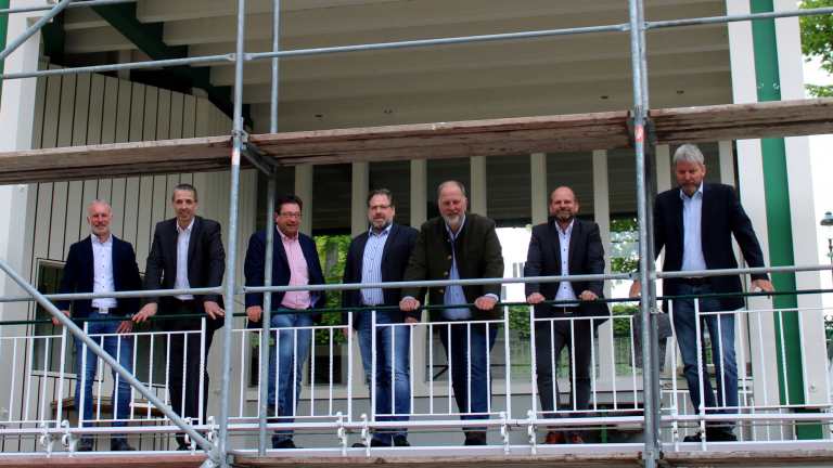 Noch steht am rechten Teil des Musikpavillons ein Gerüst. Bis zum Schützenfest sollen die Hagelschäden auf dem Dach beseitigt sein. Presseoffizier Leutnant Simon Niklas, Oberleutnant Peter Kliche, Schützenmajor Peter Liese, Schützenhauptmann Holger Harnischmacher und die Oberleutnante Frank Clemens, Andreas Roll und Christoph Schneider (v.l.) stellten in der Pressekonferenz vor dem Hochfest Neues und Bewährtes vor. Foto: mari